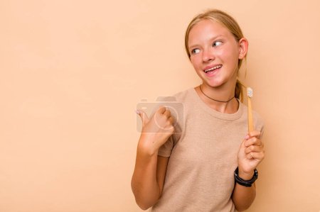 Photo for Caucasian teen girl brushing teeth isolated on beige background points with thumb finger away, laughing and carefree. - Royalty Free Image