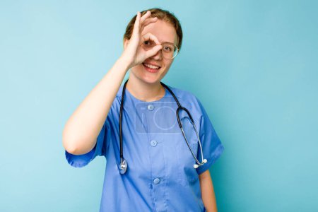 Photo for Young nurse caucasian woman isolated on blue background excited keeping ok gesture on eye. - Royalty Free Image