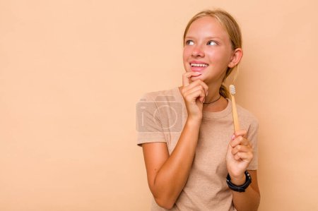 Téléchargez les photos : Caucasien adolescent fille brossage dents isolé sur beige fond détendu penser à quelque chose en regardant un copie espace. - en image libre de droit