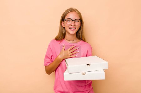Foto de Pequeña chica caucásica sosteniendo pizza aislada sobre fondo beige se ríe en voz alta manteniendo la mano en el pecho. - Imagen libre de derechos