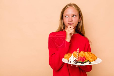 Foto de Niña caucásica sosteniendo unos waffles aislados sobre fondo beige mirando de lado con expresión dudosa y escéptica. - Imagen libre de derechos