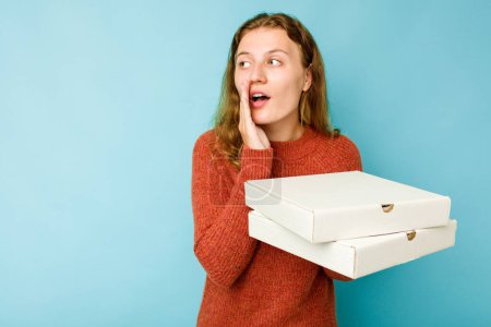 Foto de Mujer caucásica joven sosteniendo cajas de pizzas aisladas sobre fondo azul está diciendo una noticia secreta de frenado caliente y mirando a un lado - Imagen libre de derechos