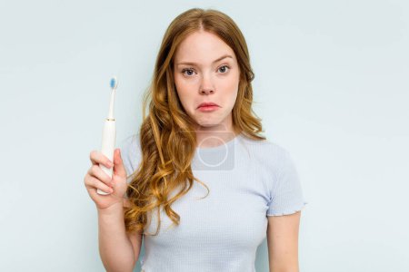 Photo for Young caucasian woman holding electric toothbrush isolated on blue background shrugs shoulders and open eyes confused. - Royalty Free Image