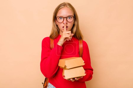 Foto de Pequeña estudiante caucásica sosteniendo hamburguesas aisladas sobre fondo beige manteniendo un secreto o pidiendo silencio. - Imagen libre de derechos