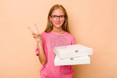 Foto de Niña caucásica sosteniendo pizza aislada sobre fondo beige mostrando el número dos con los dedos. - Imagen libre de derechos