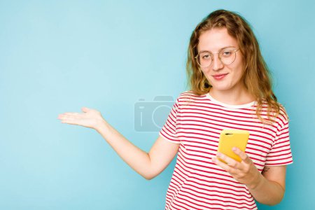 Photo for Young caucasian woman holding mobile phone isolated on blue background showing a copy space on a palm and holding another hand on waist. - Royalty Free Image