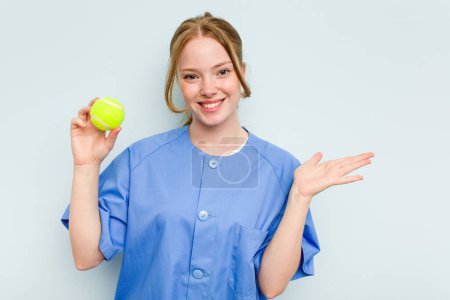 Foto de Joven fisioterapeuta caucásica sosteniendo una pelota de tenis aislada sobre fondo azul mostrando un espacio de copia en una palma y sosteniendo otra mano en la cintura. - Imagen libre de derechos