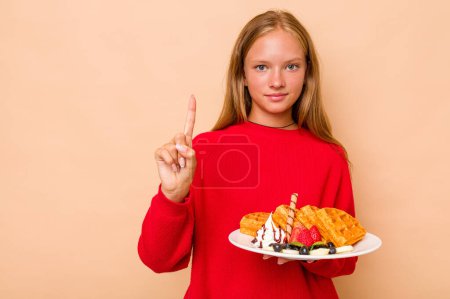 Foto de Niña caucásica sosteniendo unos waffles aislados sobre fondo beige mostrando el número uno con el dedo. - Imagen libre de derechos