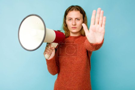 Foto de Mujer caucásica joven sosteniendo megáfono aislado sobre fondo azul de pie con la mano extendida mostrando señal de stop, lo que le impide. - Imagen libre de derechos