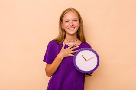 Téléchargez les photos : Petite fille caucasienne tenant une horloge isolée sur fond beige rit bruyamment en gardant la main sur la poitrine. - en image libre de droit