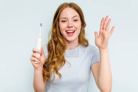 Téléchargez les photos : Jeune femme caucasienne tenant une brosse à dents électrique isolée sur fond bleu recevant une agréable surprise, excitée et levant les mains. - en image libre de droit