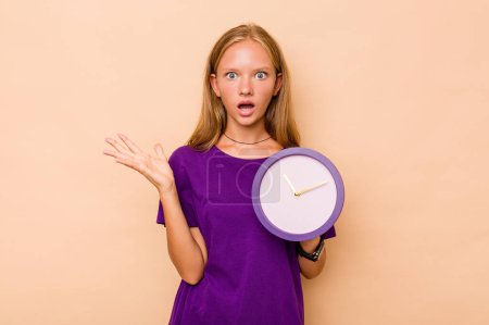 Téléchargez les photos : Petite fille caucasienne tenant une horloge isolée sur fond beige surprise et choquée. - en image libre de droit
