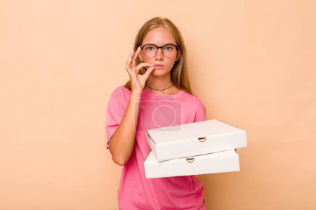 Foto de Pequeña chica caucásica sosteniendo pizza aislada sobre fondo beige con los dedos en los labios manteniendo un secreto. - Imagen libre de derechos