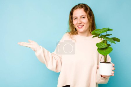 Téléchargez les photos : Jeune femme caucasienne tenant une plante isolée sur fond bleu montrant un espace de copie sur une paume et tenant une autre main à la taille. - en image libre de droit