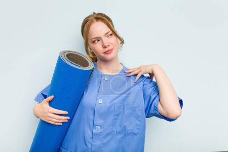 Photo for Young caucasian physiotherapist holding a mat isolated on blue background touching back of head, thinking and making a choice. - Royalty Free Image