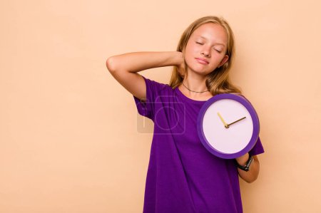 Foto de Niña caucásica sosteniendo un reloj aislado sobre fondo beige tocando la parte posterior de la cabeza, pensando y haciendo una elección. - Imagen libre de derechos
