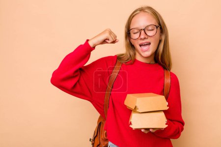Foto de Niña estudiante caucásica sosteniendo hamburguesas aisladas sobre fondo beige levantando puño después de una victoria, concepto ganador. - Imagen libre de derechos