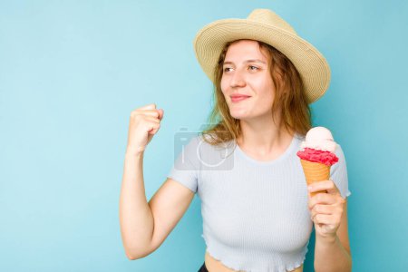 Téléchargez les photos : Jeune femme caucasienne tenant une crème glacée isolé un fond bleu levant poing après une victoire, concept gagnant. - en image libre de droit