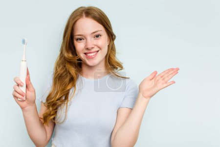 Téléchargez les photos : Jeune femme caucasienne tenant une brosse à dents électrique isolée sur fond bleu montrant un espace de copie sur une paume et tenant une autre main à la taille. - en image libre de droit