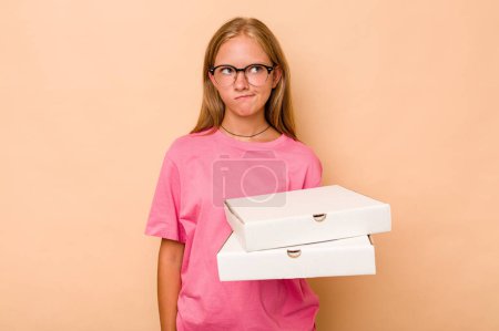 Téléchargez les photos : Petite fille caucasienne tenant pizza isolée sur fond beige confus, se sent douteuse et incertaine. - en image libre de droit