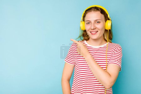Téléchargez les photos : Jeune femme caucasienne portant des écouteurs isolés sur fond bleu souriant et pointant du doigt, montrant quelque chose à l'espace vide. - en image libre de droit