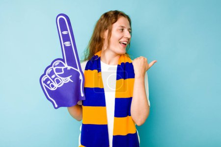 Photo for Young caucasian sports fan woman isolated on blue background points with thumb finger away, laughing and carefree. - Royalty Free Image