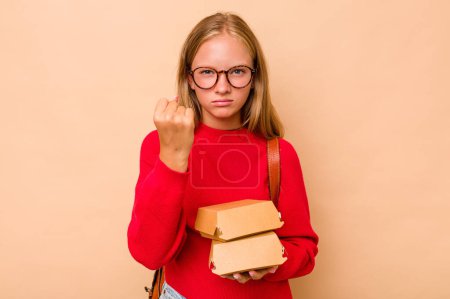 Foto de Niña estudiante caucásica sosteniendo hamburguesas aisladas sobre fondo beige mostrando puño a cámara, expresión facial agresiva. - Imagen libre de derechos