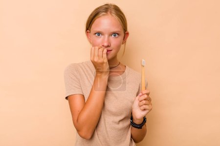 Foto de Caucásico adolescente chica cepillarse los dientes aislado en beige fondo morder las uñas, nervioso y muy ansioso. - Imagen libre de derechos