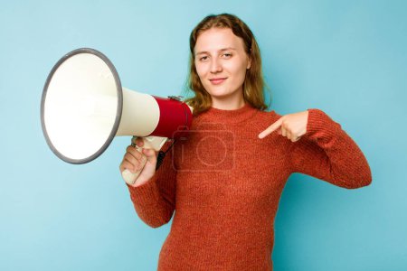 Téléchargez les photos : Jeune femme caucasienne tenant mégaphone isolé sur fond bleu personne pointant à la main vers un espace de copie de chemise, fière et confiante - en image libre de droit