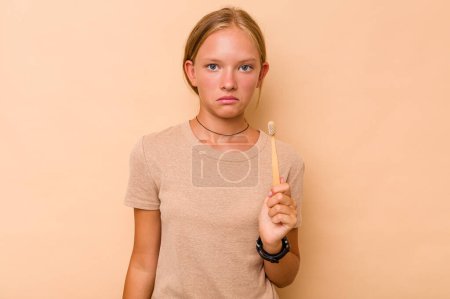 Foto de Caucásico adolescente chica cepillarse los dientes aislado en beige fondo encoge hombros y abrir los ojos confundido. - Imagen libre de derechos