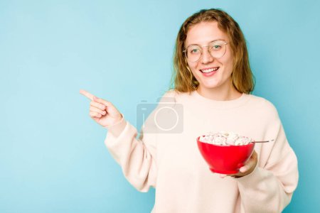 Foto de Mujer caucásica joven sosteniendo un tazón de cereales aislado sobre fondo azul sonriendo y señalando a un lado, mostrando algo en espacio en blanco. - Imagen libre de derechos