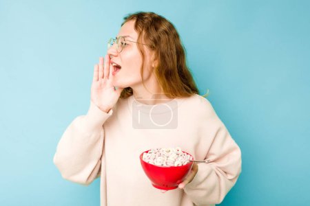 Photo for Young caucasian woman holding a bowl of cereals isolated on blue background shouting and holding palm near opened mouth. - Royalty Free Image