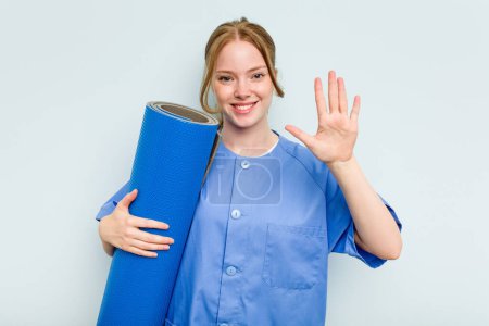 Foto de Joven fisioterapeuta caucásica sosteniendo una esterilla aislada sobre fondo azul sonriente alegre mostrando el número cinco con los dedos. - Imagen libre de derechos