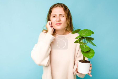 Téléchargez les photos : Jeune femme caucasienne tenant une plante isolée sur fond bleu couvrant les oreilles avec les mains. - en image libre de droit