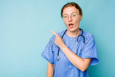 Foto de Joven enfermera caucásica aislada sobre fondo azul apuntando hacia un lado - Imagen libre de derechos