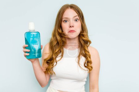 Photo for Young caucasian woman holding a mouthwash isolated on blue background shrugs shoulders and open eyes confused. - Royalty Free Image