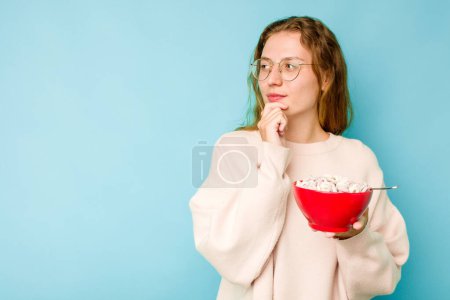 Téléchargez les photos : Jeune femme caucasienne tenant un bol de céréales isolé sur fond bleu regardant de côté avec une expression douteuse et sceptique. - en image libre de droit