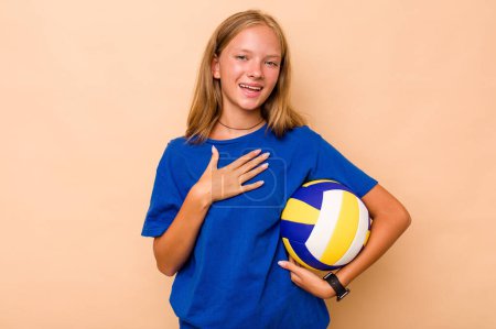 Téléchargez les photos : Petite fille caucasienne jouant au volley isolé sur fond beige rit bruyamment en gardant la main sur la poitrine. - en image libre de droit