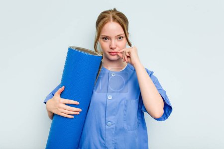 Photo for Young caucasian physiotherapist holding a mat isolated on blue background with fingers on lips keeping a secret. - Royalty Free Image