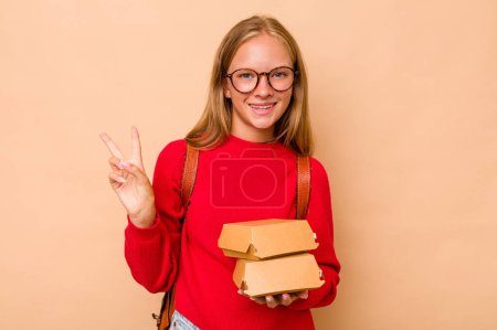 Téléchargez les photos : Petite étudiante caucasienne tenant des hamburgers isolés sur fond beige joyeuse et insouciante montrant un symbole de paix avec les doigts. - en image libre de droit