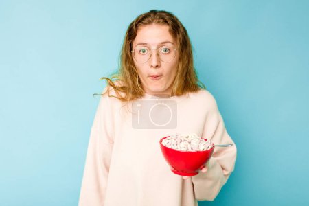 Photo for Young caucasian woman holding a bowl of cereals isolated on blue background shrugs shoulders and open eyes confused. - Royalty Free Image