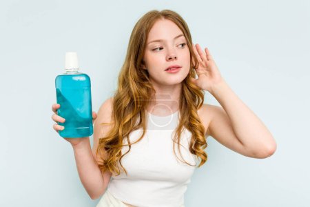 Photo for Young caucasian woman holding a mouthwash isolated on blue background trying to listening a gossip. - Royalty Free Image