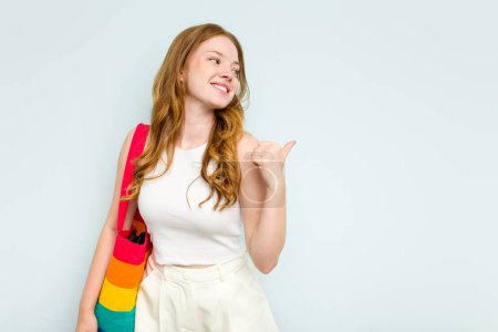 Foto de Mujer caucásica joven sosteniendo LGTBI bolsa aislada en puntos de fondo azul con el dedo pulgar lejos, riendo y sin preocupaciones. - Imagen libre de derechos