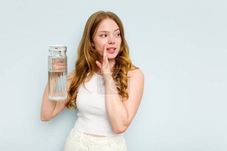 Téléchargez les photos : Jeune femme caucasienne tenant pot d'eau isolé sur fond bleu dit une nouvelle secrète de freinage chaud et regardant de côté - en image libre de droit