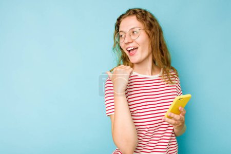 Photo for Young caucasian woman holding mobile phone isolated on blue background points with thumb finger away, laughing and carefree. - Royalty Free Image