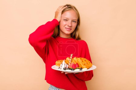 Foto de Pequeña chica caucásica sosteniendo un waffles aislado sobre fondo beige siendo sorprendido, ella ha recordado reunión importante. - Imagen libre de derechos