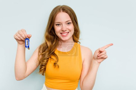 Foto de Young caucasian woman holding home keys isolated on blue background smiling and pointing aside, showing something at blank space. - Imagen libre de derechos