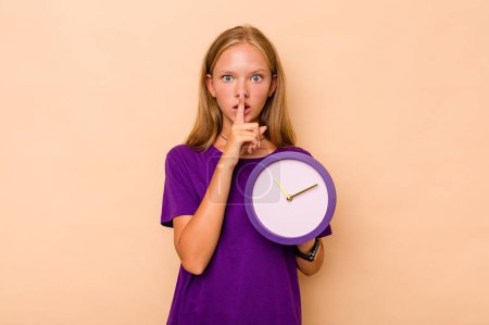 Téléchargez les photos : Petite fille caucasienne tenant une horloge isolée sur fond beige gardant un secret ou demandant le silence. - en image libre de droit