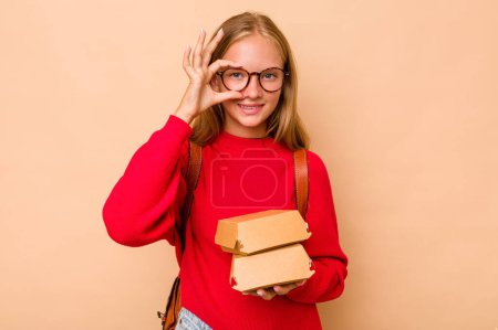 Foto de Pequeña chica estudiante caucásica sosteniendo hamburguesas aisladas sobre fondo beige excitado manteniendo el gesto bien en el ojo. - Imagen libre de derechos