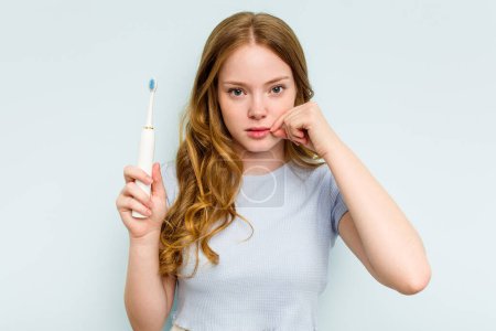 Téléchargez les photos : Jeune femme caucasienne tenant une brosse à dents électrique isolée sur fond bleu avec les doigts sur les lèvres gardant un secret. - en image libre de droit
